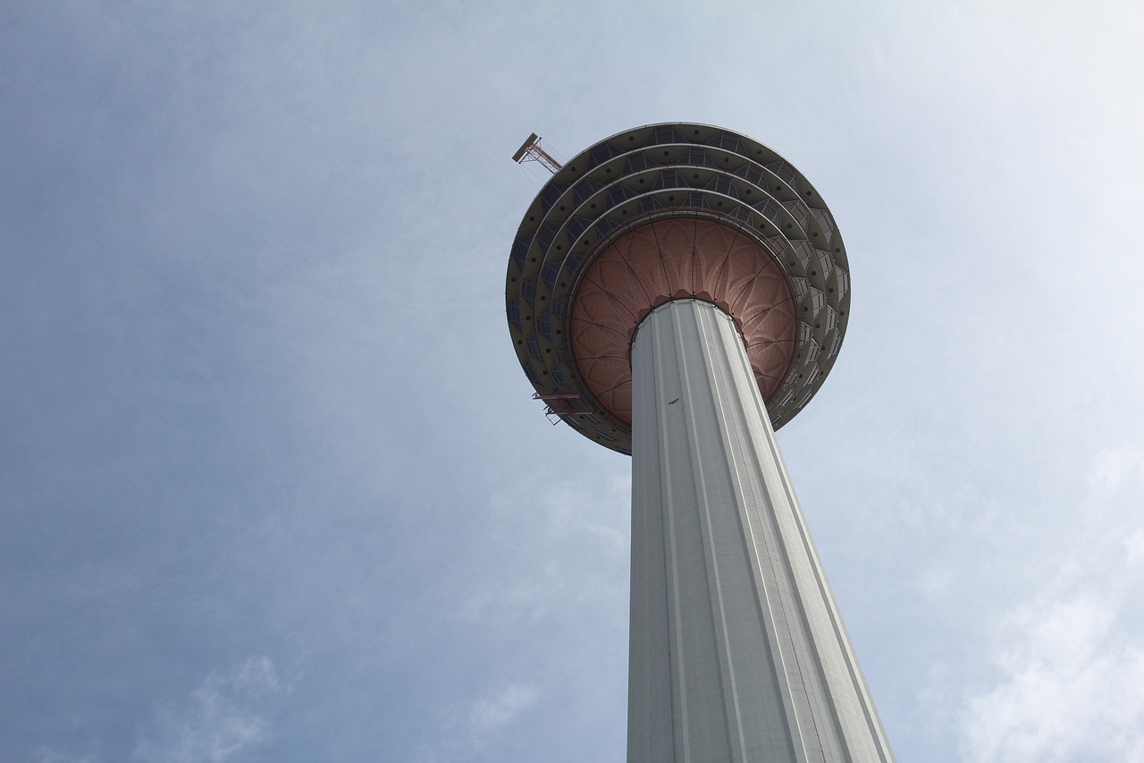 Looking up at a Malaysian telecommunications and observation tower, its viewing pod ringed against a blue sky.