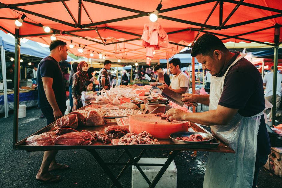 Stalls and shoppers at a bustling Malaysian pasar malam, tables piled high under glowing red canopies.