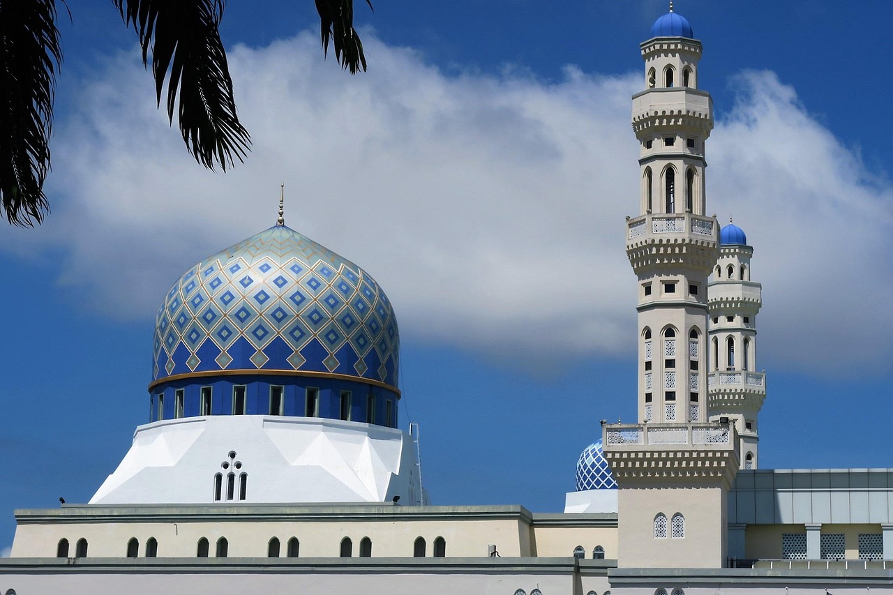 A grand Malaysian mosque with patterned blue domes and tiered minarets against a tropical sky.