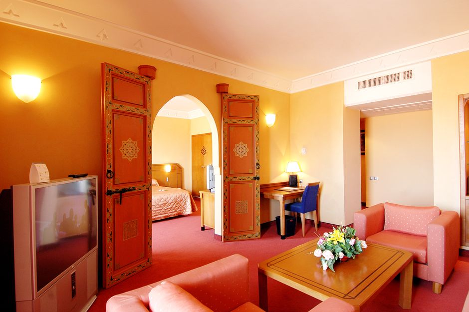 A suite with a separate sitting area in warm ochre and oxblood tones, an arched doorway through to the bedroom and a flat-screen TV.
